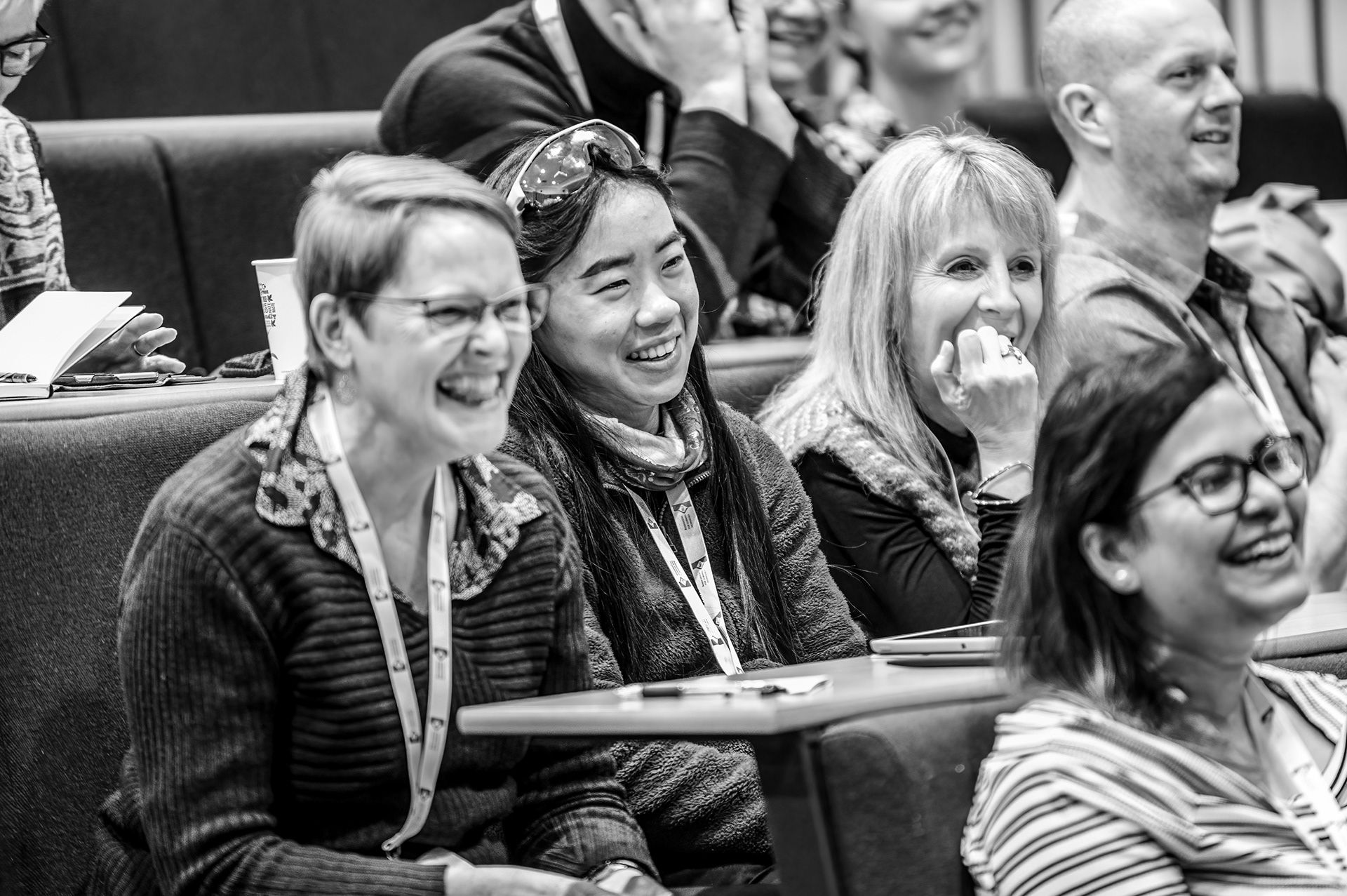 People sitting at desks in a lecture hall, watching a presentation and smiling.